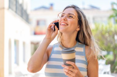Açık havada cep telefonu kullanan ve elinde mutlu bir ifadeyle kahve tutan genç ve güzel bir kadın.