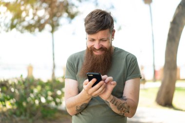 Kızıl saçlı sakallı adam cep telefonuyla mesaj yolluyor.