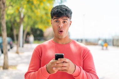 Dışarıdaki İspanyol genç adam kameraya bakarken şaşırmış bir ifadeyle cep telefonunu kullanıyor.