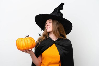 Beyaz arka planda izole edilmiş balkabağı tutan cadı kostümü giyip yan tarafa bakıyor ve gülümsüyor.