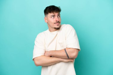 Beyaz tenli yakışıklı adam mavi arka planda izole edilmiş. Omuzlarını kaldırırken şüpheyle hareket ediyor.