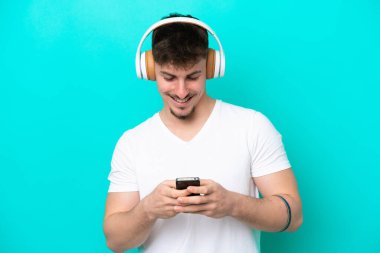 Beyaz tenli yakışıklı adam mavi arka planda izole edilmiş müzik dinliyor ve cep telefonu arıyor.