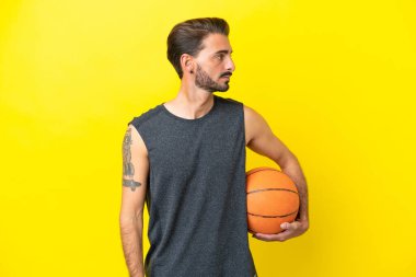 Sarı arka planda izole edilmiş yakışıklı genç basketbolcu yan tarafa bakıyor.