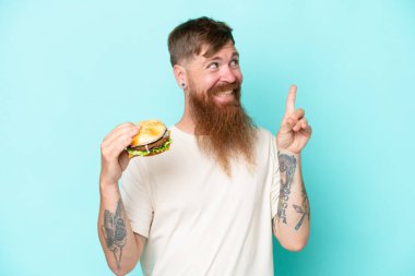 Uzun sakallı kızıl saçlı bir adam mavi arka planda izole edilmiş bir burger tutuyor. Çözümü bulmak için parmağını kaldırıyor.