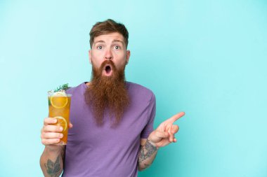 Uzun sakallı kızıl saçlı adam mavi arka planda izole edilmiş bir kokteyl tutuyor. Şaşırmış ve işaret ediyor.
