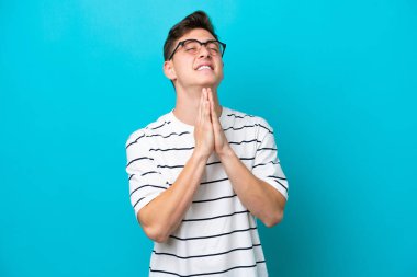 Young handsome Brazilian man isolated on blue background keeps palm together. Person asks for something