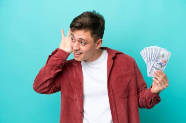 Genç Brezilyalı bir adam mavi arka planda izole edilmiş bir sürü para alıyor. Kulağına elini sürerek bir şeyler dinliyor.