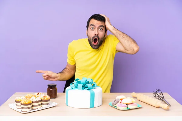 Man in a table with a big cake surprised and pointing finger to the side