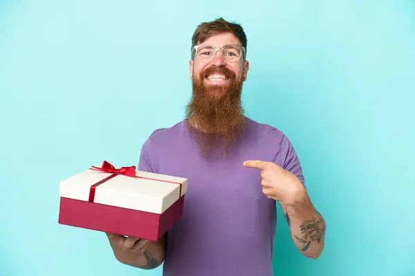 Redhead man with long beard holding a gift isolated on blue background with surprise facial expression