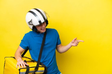 Sırt çantalı genç Brezilyalı adam sarı arka planda izole edilmiş, parmağını yana doğru uzattı ve bir ürün sundu.