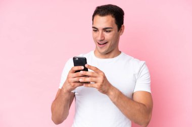 Young caucasian man isolated on pink background looking at the camera while using the mobile with surprised expression