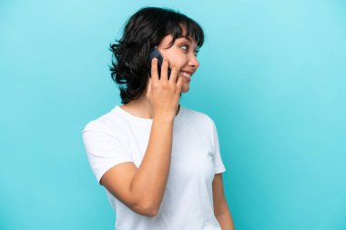 Arjantinli genç bir kadın mavi arka planda izole edilmiş cep telefonuyla biriyle sohbet ediyor.