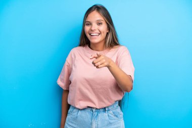 Mavi arka planda izole edilmiş genç beyaz kadın şaşırmış ve ön tarafı işaret ediyor.