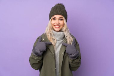 Kışlık şapkalı, mor arka planda, sürpriz bir yüz ifadesi olan genç sarışın kız.