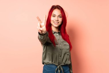 Genç kızıl saçlı kız pembe arka planda izole edilmiş gülümsüyor ve zafer işareti gösteriyor.