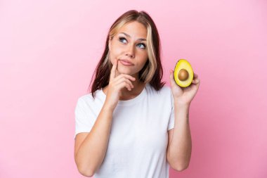 Avokado tutan genç Rus kadın pembe arka planda izole edilmiş, yukarı bakarken şüpheleri var.