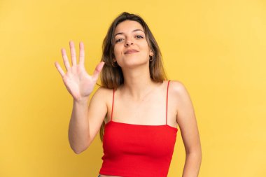 Sarı arka planda izole edilmiş genç beyaz kadın parmaklarıyla beşi sayıyor.