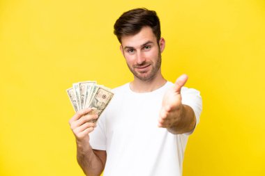 Kafkasyalı genç adam sarı arka planda izole edilmiş bir sürü para alıyor ve iyi bir anlaşma için el sıkışıyor.