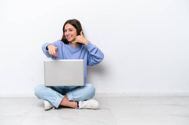 Yerde dizüstü bilgisayarlı beyaz bir kadın oturuyor. Beyaz arka planda izole edilmiş. Telefon hareketleriyle ön tarafı işaret ediyor.