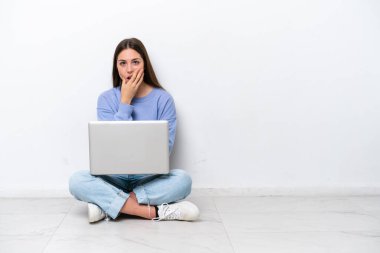Yerde dizüstü bilgisayarlı beyaz bir kadın oturuyor. Beyaz arka planda izole edilmiş. Şaşırmış ve şoka uğramış.
