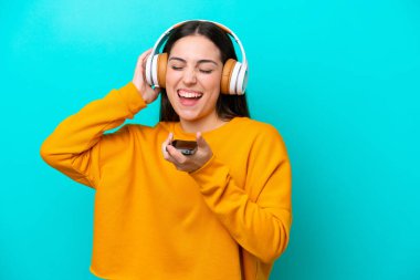 Mavi arka planda izole edilmiş genç bir kız cep telefonuyla müzik dinliyor ve şarkı söylüyor.