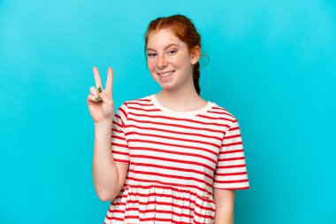 Mavi arka planda izole edilmiş genç, kırmızı bir kadın gülümsüyor ve zafer işareti gösteriyor.