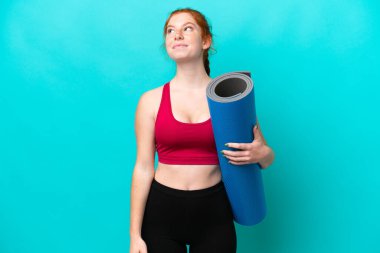 Genç spor kırmızısı kadın yoga derslerine giderken elinde bir minder, mavi arka planda izole bir şekilde yukarı bakıyor.