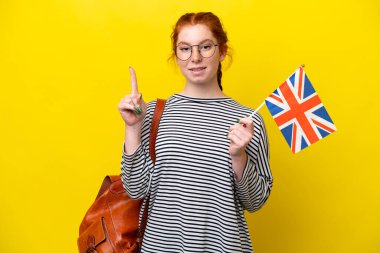 Birleşik Krallık bayrağını tutan genç İspanyol kadın sarı arka planda izole edilmiş bir şekilde parmağını kaldıran bir fikir düşünüyor.