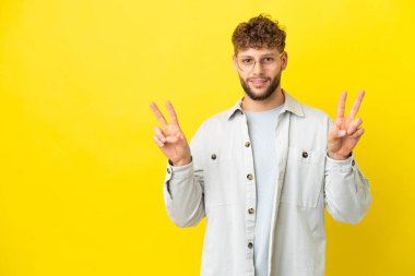 Sarı arka planda izole edilmiş yakışıklı beyaz adam iki eliyle zafer işareti gösteriyor.