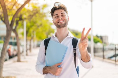 Açık havada gülümseyen ve zafer işareti gösteren yakışıklı bir öğrenci.
