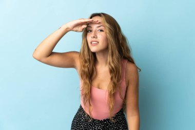 Mavi arka planda izole edilmiş genç sarışın bir kadın bir şeye bakmak için elini uzaklara bakıyor.