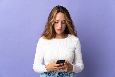 Young blonde woman isolated on purple background using mobile phone