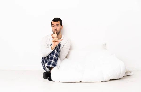 Caucasian man in pajamas sitting on the floor at indoors pointing to the side and doing silence gesture