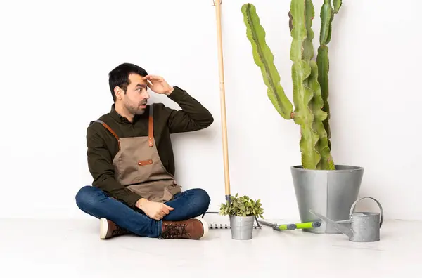 Gardener man sitting on the floor at indoors with surprise expression while looking side