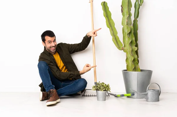 Gardener man sitting on the floor at indoors pointing finger to the side and presenting a product