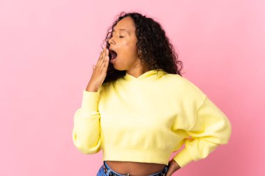 Kübalı genç bir kız pembe arka planda izole edilmiş esniyor ve ağzını eliyle açık tutuyor.