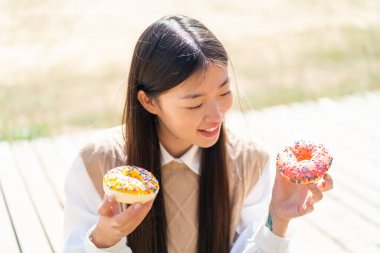 Genç Çinli bir kadın açık havada elinde mutlu bir ifadeyle donut tutuyor.