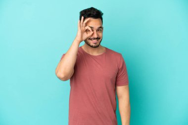 Mavi arka planda izole edilmiş genç beyaz adam parmaklarıyla tamam işareti gösteriyor.