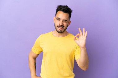 Mor arka planda izole edilmiş genç beyaz adam parmaklarıyla tamam işareti gösteriyor.