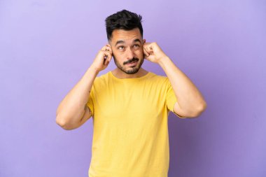 Mor arka planda izole edilmiş genç beyaz adam hayal kırıklığına uğramış ve kulaklarını kapatmış.