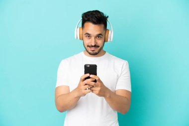 Kafkasyalı genç adam mavi arka planda izole edilmiş. Cep telefonuyla müzik dinliyor ve ön tarafa bakıyor.