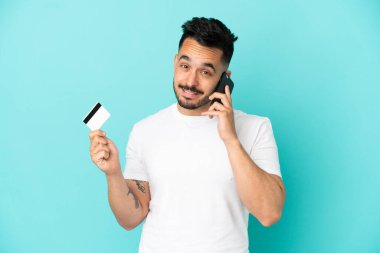 Kafkasyalı genç adam mavi arka planda izole edilmiş cep telefonuyla sohbet ediyor ve elinde bir kredi kartı tutuyor.