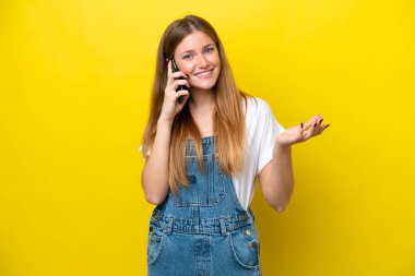 Sarı arka planda izole edilmiş genç beyaz bir kadın cep telefonuyla sohbet ediyor.