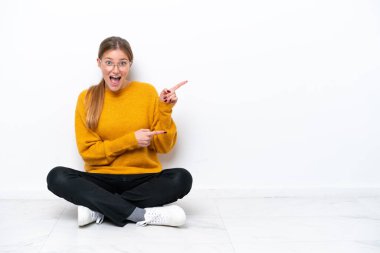 Beyaz arka planda izole bir şekilde yerde oturan genç beyaz kadın şaşırmış ve işaret ediyor.