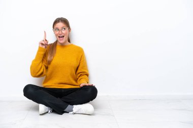 Beyaz arka planda izole bir şekilde yerde oturan beyaz tenli genç kadın çözümün farkına varmak için parmağını kaldırıyor.