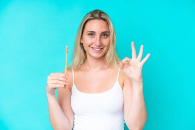 Mavi arka planda dişlerini fırçalayan genç beyaz kadın parmaklarıyla işaret veriyor.