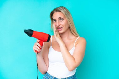 Kafkasyalı genç bir kadın elinde saç kurutma makinesiyle mavi arka planda bir şeyler fısıldıyor.