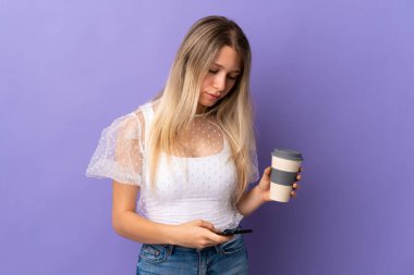 Mor arka planda izole edilmiş genç sarışın kadın elinde kahve ve bir cep telefonu tutuyor.