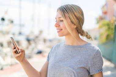 Açık havada cep telefonu kullanan genç sarışın kadın. Mutlu bir ifadeyle.