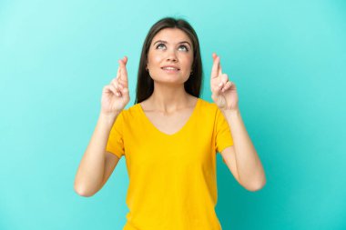 Mavi arka planda izole edilmiş genç beyaz kadın parmaklarını çapraz yapıp en iyisini diliyor.